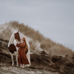 Junge Frau am Strand in den Dünen mit braun weißem Pferd. Sie trägt ein rotes Kleid. Pferdefotografie